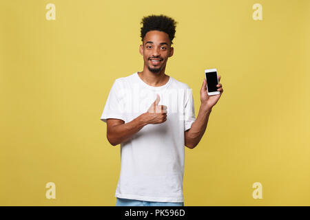 Portrait of attractive young black man isolated on yellow background, holding blank smartphone, smiling at camera, showing thubms up gesture, feeling happy. Stock Photo