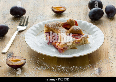 Piece of homemade plum pie on a white plate with organic plums and fork on wooden table. Closeup Stock Photo