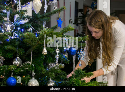 Christmas tree being decorated by a young girl with red hair. Blue and silver decorations. Baubles, beads and icicles. Stock Photo