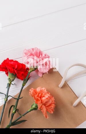 Three multicolored carnation flowers and shopping bag. White wooden surface background. Top view. Stock Photo