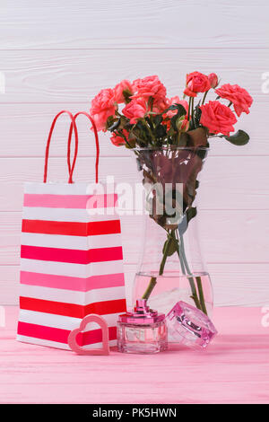 Gifts for valentines day. Shopping bag, rose flowers in a vase and perfume bottle. Pink wooden background. Stock Photo