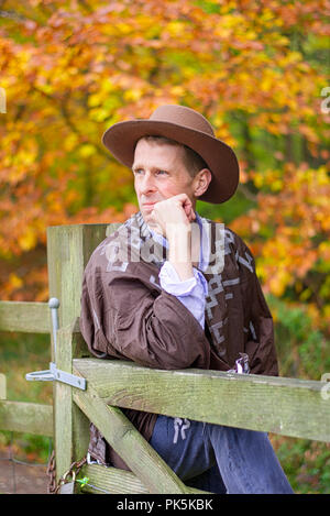 A middle aged man wearing a cowboy hat reclines against a fence post. Stock Photo