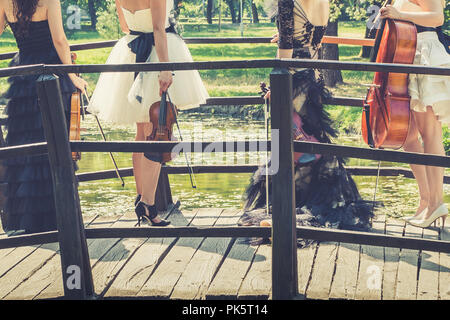 Music and nature concept. Female musical quartet with string instruments, one cello and three violins, standing on bridge, prepares to play in nature  Stock Photo