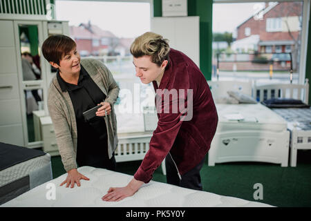 A saleswoman is assisting a young man as he shops for a new matress. She is talking to him while he tests a display. Stock Photo