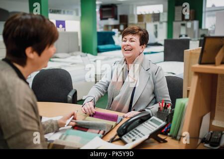 Mature woman is laughing with the sales clerk while looking at fabric swatches in a furniture shop. Stock Photo