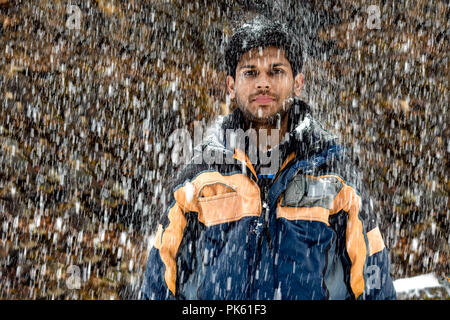 Young, Fair and Handsome Indian boy in early 20s in skiing costume, wearing snowsuit, standing still and posing in neutral mood during heavy snowfall Stock Photo