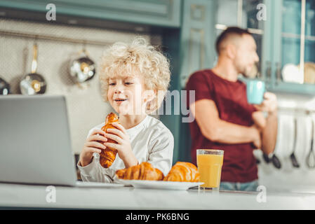 Cute blonde-haired boy eating croissant and watching cartoons Stock Photo