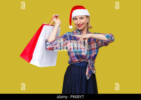 Cheerful modern middle aged woman in red santa cap and checkered shirt standing and pointing finger to shopping bags and toothy smile, looking at came Stock Photo