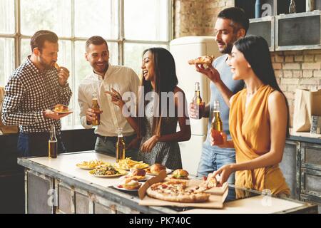 Home party. Full length of cheerful young people enjoying home party while communicating and eating snacks on the kitchen. Stock Photo