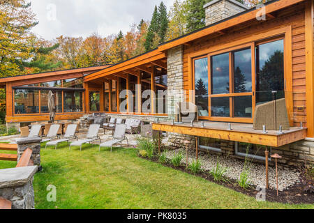 Rear view of luxurious stained cedar and timber wood home with panoramic windows, deck and natural stone patio in autumn, Quebec Stock Photo