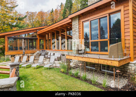 Rear view of luxurious stained cedar and timber wood home with panoramic windows, deck and natural stone patio in autumn, Quebec Stock Photo