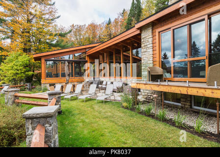 Rear view of luxurious stained cedar and timber wood home with panoramic windows, deck and natural stone patio in autumn, Quebec Stock Photo