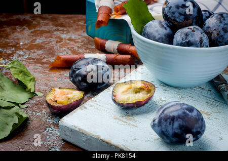 Fresh Purple Plums in a pottery blue bowl   one plum is halved and the bright fruit pulp glows on dark brown rustic background, selective focus, Copy  Stock Photo