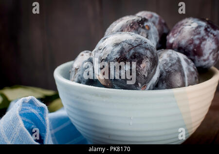 Blue Plums in a pottery bowl and leaves on dark brown rustic background, selective focus, Copy Space Stock Photo