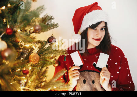 beautiful girl holding two credit cards for discount and buying gifts on background of golden christmas tree with lights and presents in festive room. Stock Photo