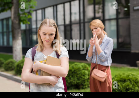 disappointed teen daughter with books looking down while her mother smoking cigarette blurred on background Stock Photo