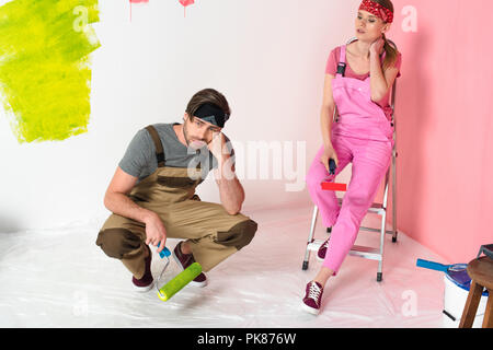 tired young couple in working overalls sitting with paint rollers Stock Photo