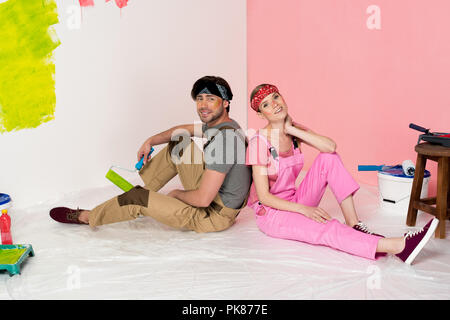 tired young couple in working overalls sitting on floor surrounded by painting tools Stock Photo