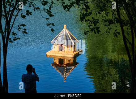 Man photographing small wooden birdhouse on his phone with a beautiful paintings in the middle of a lake surrounded by tree branches and leaves Stock Photo