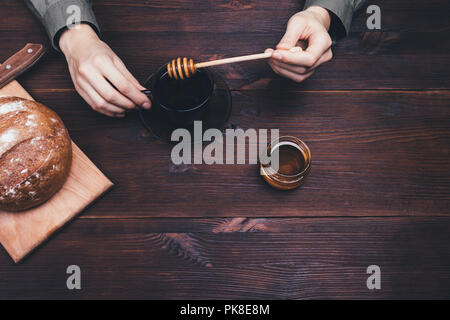 Woman adding honey to cup of tea or coffee next to round loaf of rye bread on a wooden table, top view. Stock Photo