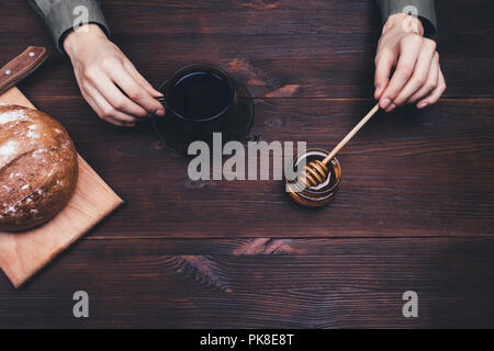 Simple rural breakfast with tea, honey and rye bread. Woman's hands holding spoon and cup on dark wooden table, top view. Stock Photo