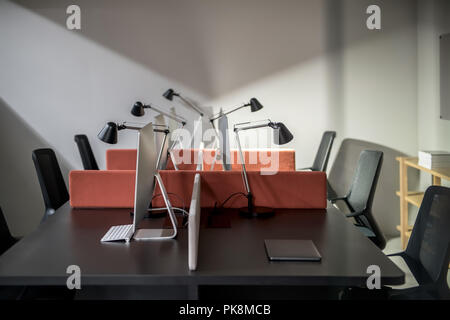 Workplaces in an ofice with gray walls. There are dark tables with peach partitions and computers, black chairs, wooden shelves with books, luminous l Stock Photo