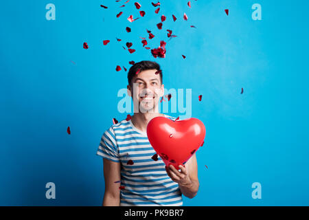 A young man in love holding red heart, confetti falling on him. Stock Photo