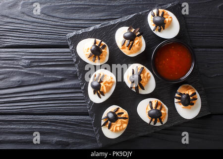 Festive Halloween appetizer: halved deviled eggs decorated with olive spiders close-up served with ketchup on a black table. horizontal top view from  Stock Photo