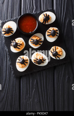 Halves deviled eggs decorated with olive spiders close-up served with ketchup on a black table. Halloween food. Vertical top view from above Stock Photo