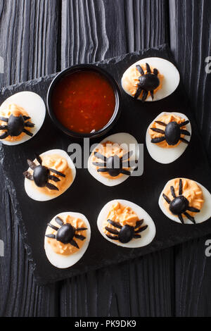 Halloween appetizer deviled eggs stuffed with mustard and decorated with olive spiders and cobweb close-up on a blackboard. Vertical top view from abo Stock Photo