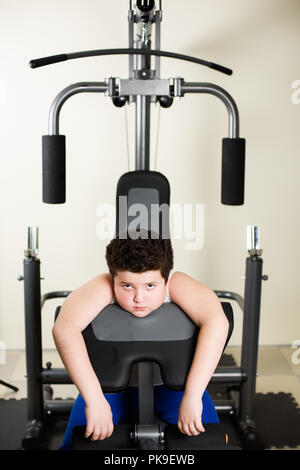 tired thick small kid sitting on training machine in gym indoors Stock Photo