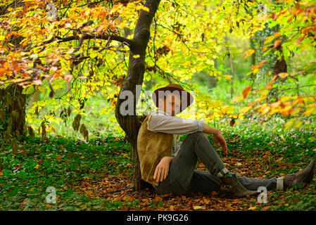 A man dressed like a cowboy sits on the floor leaning against a small tree trunk Stock Photo