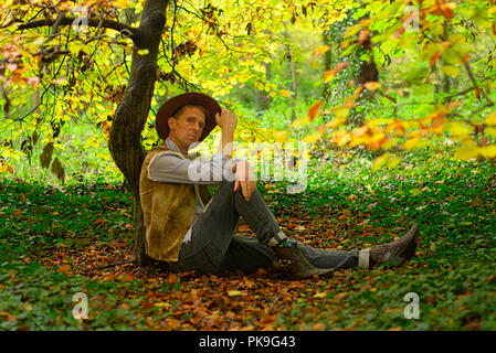 A man dressed like a cowboy sits on the floor leaning against a small tree trunk Stock Photo