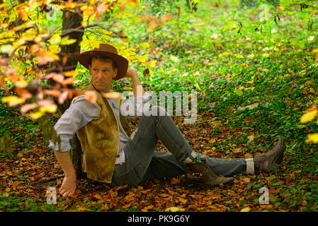 A man dressed like a cowboy sits on the floor leaning against a small tree trunk Stock Photo