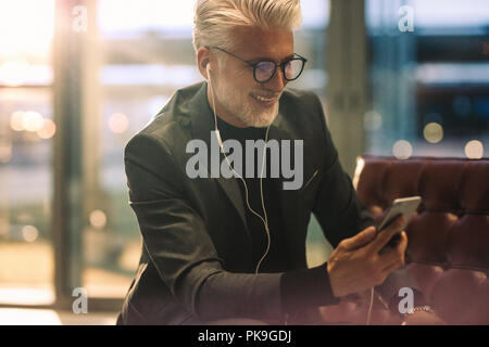Mature businessman sitting in office lobby making video call with his smart phone. Caucasian man with white hair in office making video call. Stock Photo