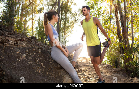 Fit young woman in sportswear sitting on the rock and relaxing after trail run training with male stretching leg. Man and woman trail runner taking br Stock Photo