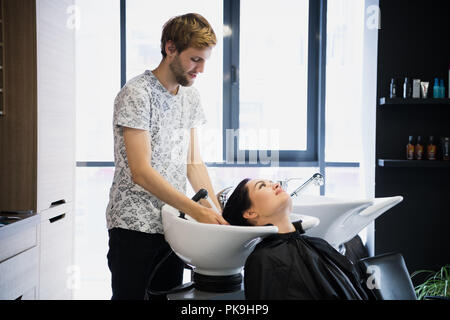 Young hipster male hairdresser washing hair of a beautiful brunette female woman client in a saloon. Stock Photo
