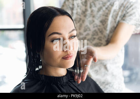 Confident young woman is looking at her reflection while hairdresser combing and cutting client's hair Stock Photo