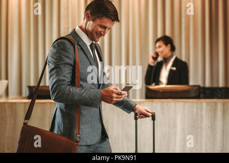 Businessman walking in hotel lobby with suitcase and using his smart phone. Male business traveler in hotel hallway with cellphone and luggage. Stock Photo