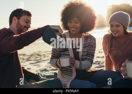 Man pouring coffee in woman's cup at beach. Group of young friends having coffee at beach. Stock Photo