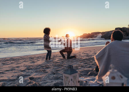 Young man kneeling and proposing woman with a ring at beach. Beautiful view of couple proposing at beach sunset with people sitting in front wrapped i Stock Photo