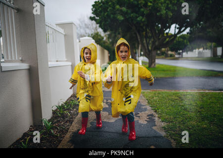 Happy young girls in raincoat and rubber boots running outdoors. Twin sisters in waterproof coats in a rainy day. Stock Photo
