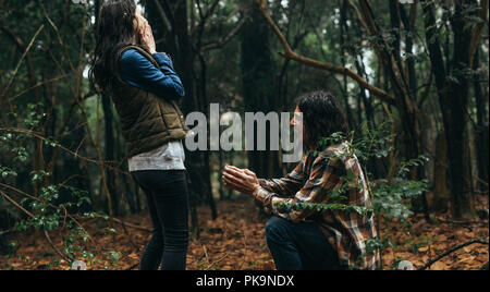 Couple in forest, with man proposing his girlfriend. Man kneeling holding a ring and making a marriage proposal to woman with surprised expression. Stock Photo