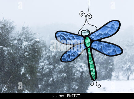 Cold snowy day with oak trees seen through a window with bright blue and green stained-glass dragonfly sun catcher hanging in window Stock Photo