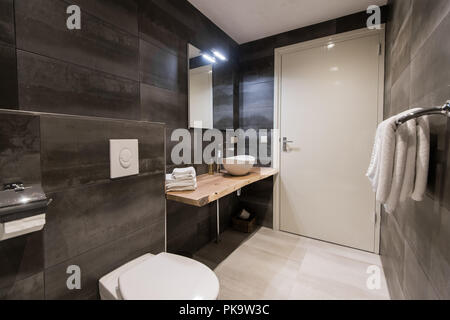 Contemporary bathroom tiled with the white and gray tiles. There is a white sink with accessories, big mirror, towel holder, toilet, white stand with a towel and a book. Horizontal. Stock Photo
