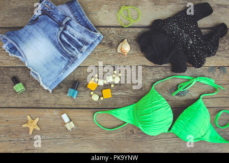 Summer women's clothing and accessories: bathing suit, denim shorts, t-shirt, shells, nail polish on old wooden background. Stock Photo