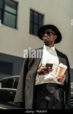 Fashionable african american businessman wearing fedora hat using smartphone and holding coffee cup Stock Photo