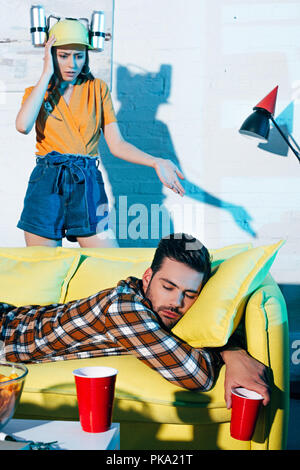 shocked girl in beer hat looking at drunk man sleeping on couch after home party Stock Photo