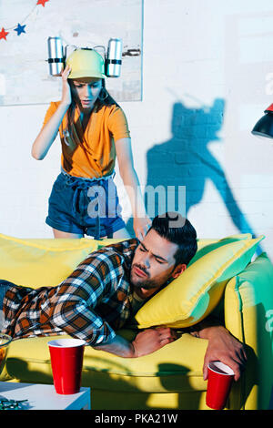 shocked girl in beer hat looking at drunk man lying on sofa after home party Stock Photo