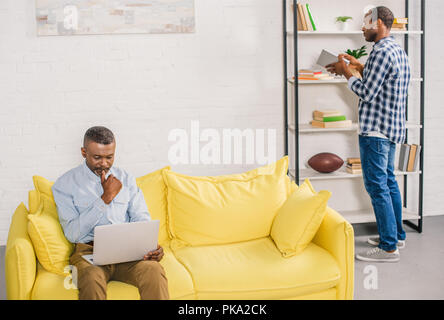 senior man using laptop while adult son holding book near bookshelves at home Stock Photo